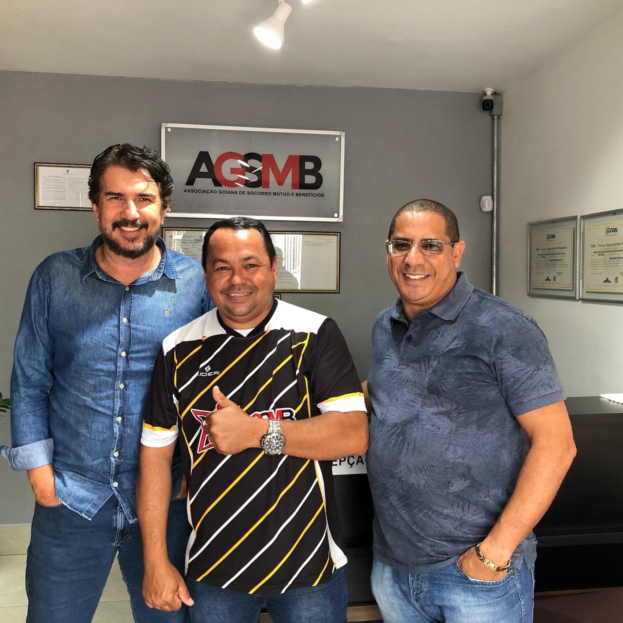 Três homens posam na sede da AGSMB diante da placa da associação